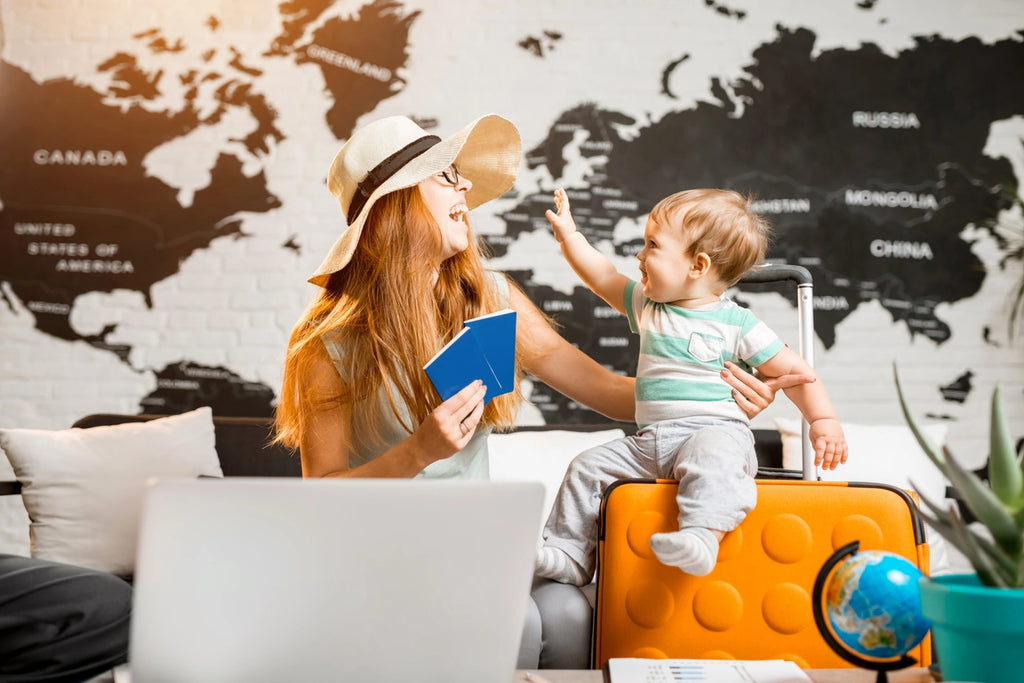Mom traveling with a baby holding passports and laughing at her baby sitting on a suitcase in front of a world map